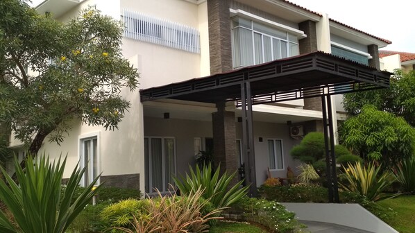 A modern two-story house with a covered patio area, surrounded by lush greenery and a variety of plants.