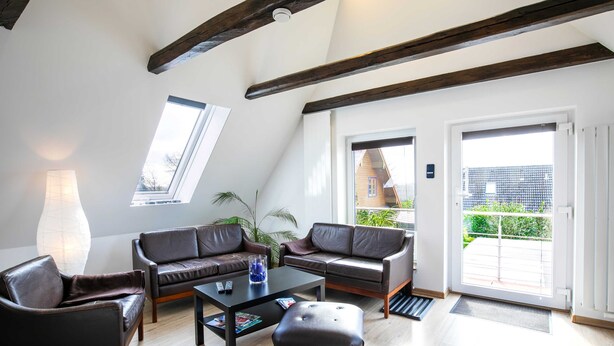 Ein modernes Wohnzimmer mit hoher Decke, Holzbalken, zwei Ledersofas, einem Couchtisch und einem großen Fenster.