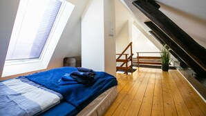 Ein Schlafzimmer mit Dachfenster, Holzboden, einem Bett mit blauer Bettwäsche, einer Treppe und einem Blumentopf.