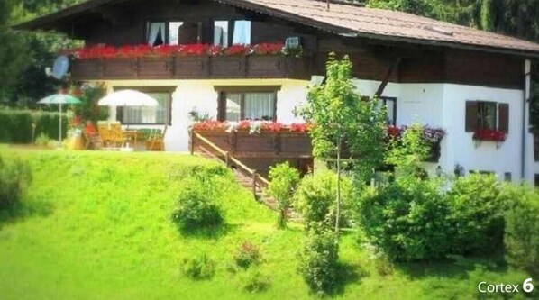 A two-story chalet with a balcony, surrounded by lush greenery.