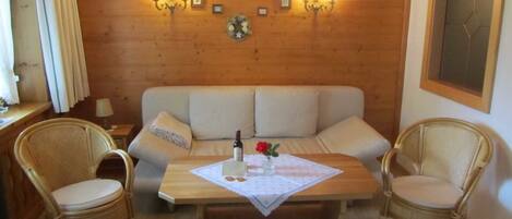 Une pièce chaleureuse avec un mur en bois, un canapé, deux fauteuils en osier, une table avec un set de table et des lampes fixées au mur.