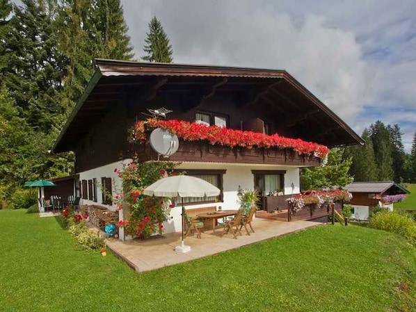 A chalet-style building with a sloped roof, a balcony with red flowers, and a patio area with outdoor furniture.