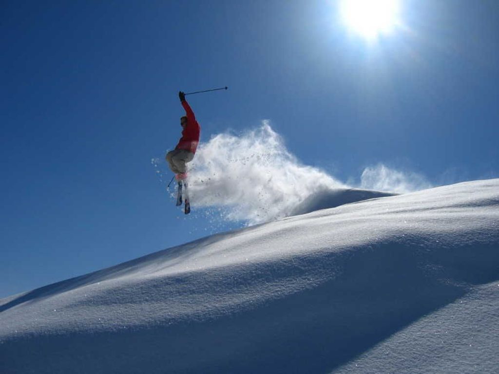 Une personne fait du ski sur une pente enneigée, avec un ciel bleu dégagé et le soleil en arrière-plan.