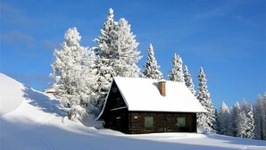 Una cabaña con techo nevado, rodeada de árboles cubiertos de nieve y un cielo azul despejado.