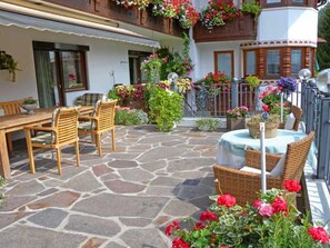 A stone patio with outdoor furniture, a balcony with flower boxes, and a building with a white facade and wooden accents. - Vacation apartment Landleben for 4-5 persons - Landhaus Dengg (Gerlos)