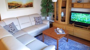 Ein modernes Wohnzimmer mit einem beigefarbenen Sofa, einem Holzschrank mit Glastüren, einem Fernseher, einem Couchtisch und einer Topfpflanze.