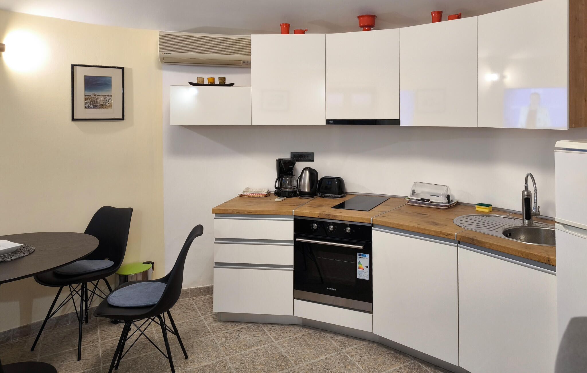 A compact kitchen with white cabinets, a built-in oven, a coffee maker, and a toaster. There is a dining table with chairs and a framed picture on the wall.
