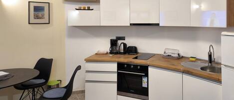 A compact kitchen with white cabinets, a built-in oven, a coffee maker, and a toaster. There is a dining table with chairs and a framed picture on the wall.