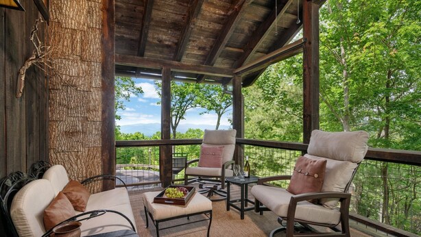 A covered outdoor seating area with cushioned chairs and a table, overlooking a forested landscape.