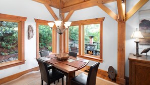 A dining area with a wooden table, chairs, and a chandelier. There are large windows with a view of greenery outside.