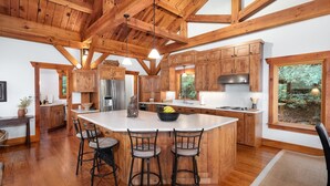 A spacious kitchen with wooden cabinets, a large island with bar stools, stainless steel appliances, and a view of greenery through a window.