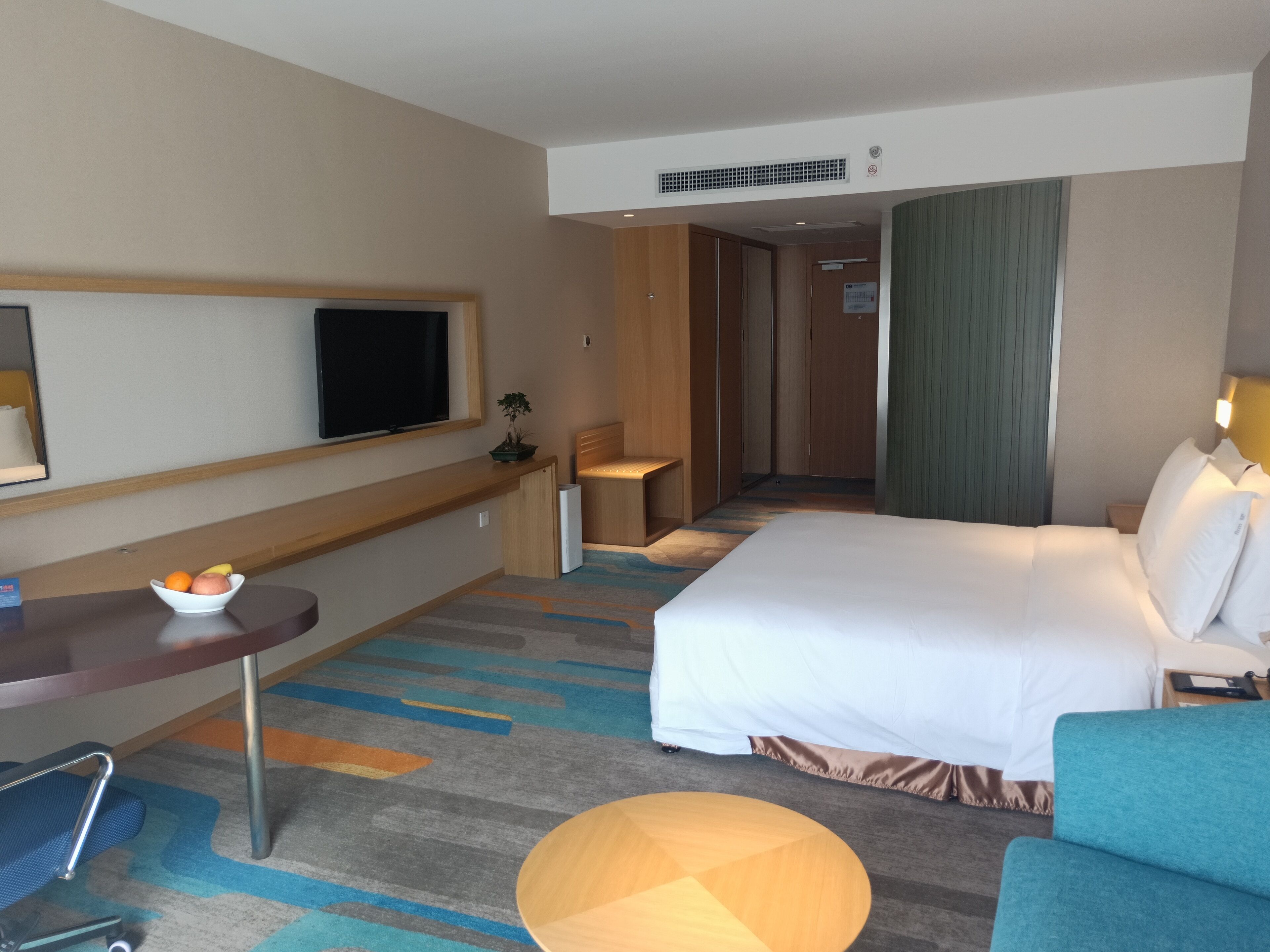 A hotel room with a bed, a desk with a chair, a television, and a small table with a bowl of fruit.