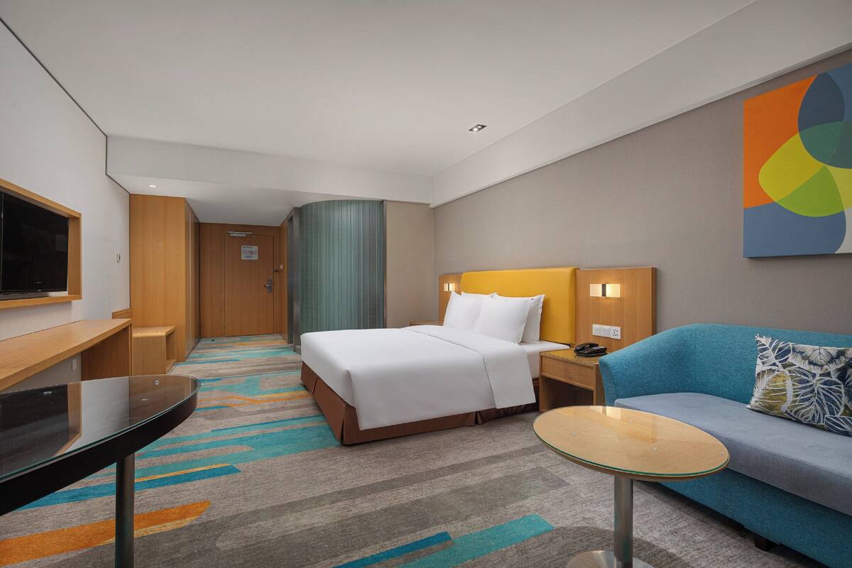 A hotel room with a bed, a sofa, a small table, a TV, and a wall-mounted artwork.