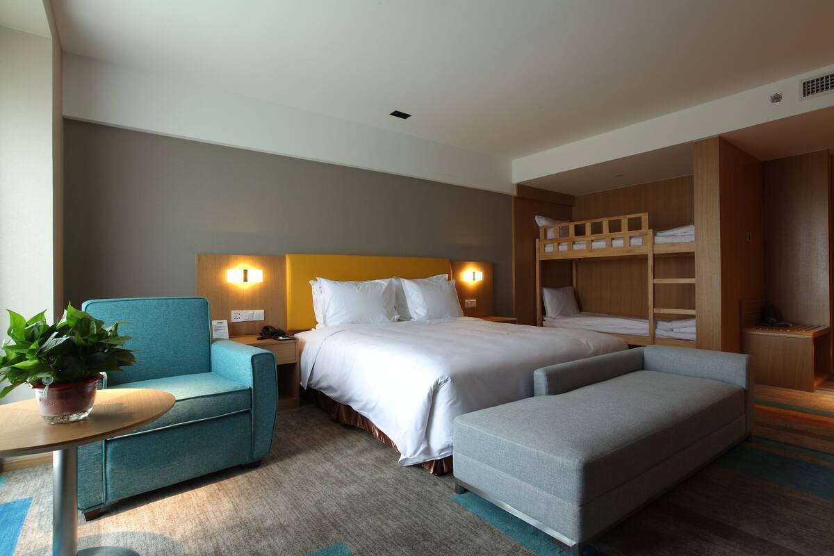 A hotel room with a bed, a sofa, a chair, a small table with a plant, and a wooden storage unit.