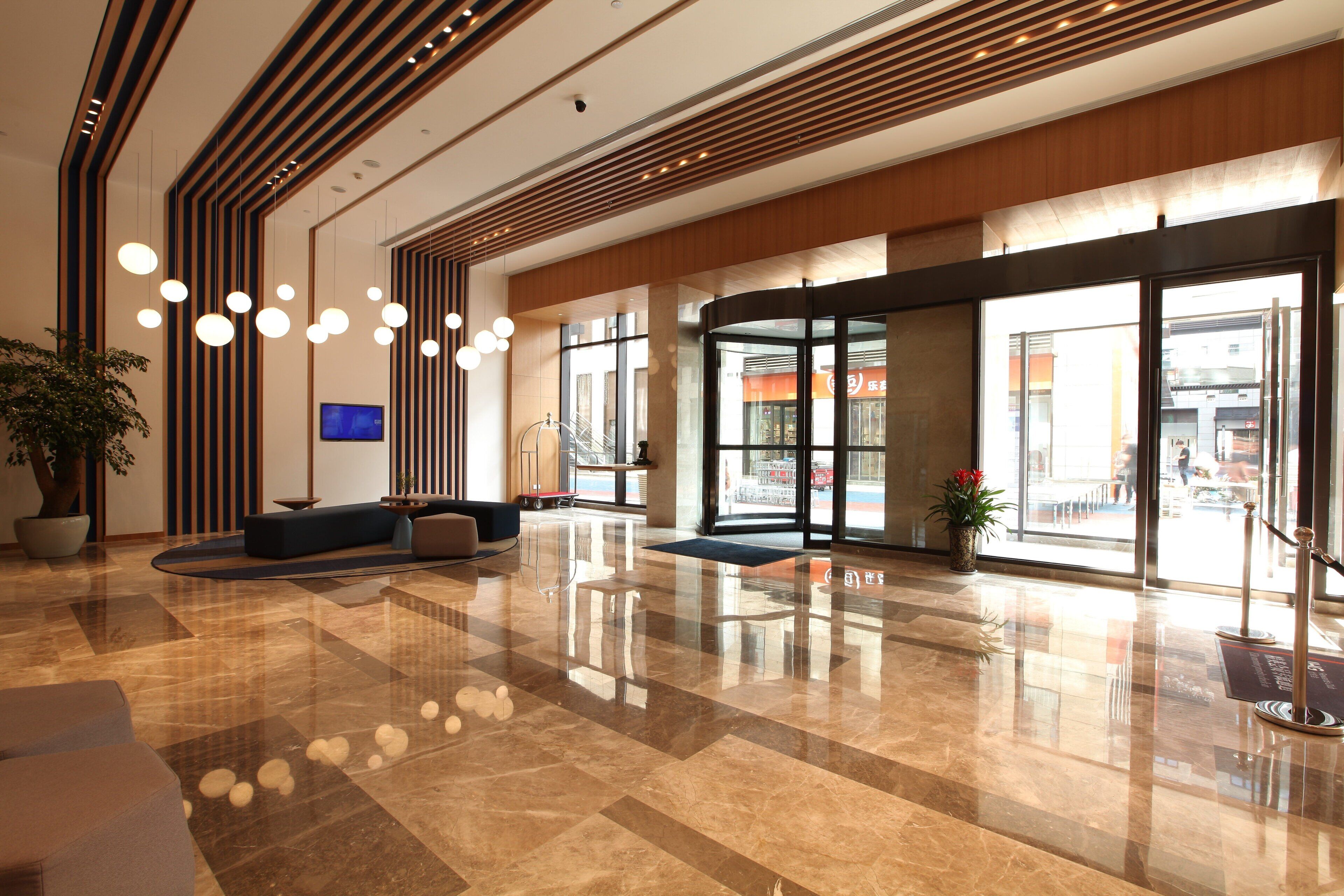 Um lobby moderno com piso de mármore polido, área de estar com sofás, uma grande planta em vaso e uma tela de televisão.