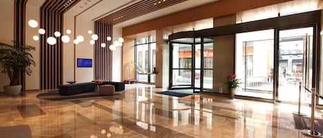 A modern lobby with a polished marble floor, a seating area with sofas, a large potted plant, and a television screen.