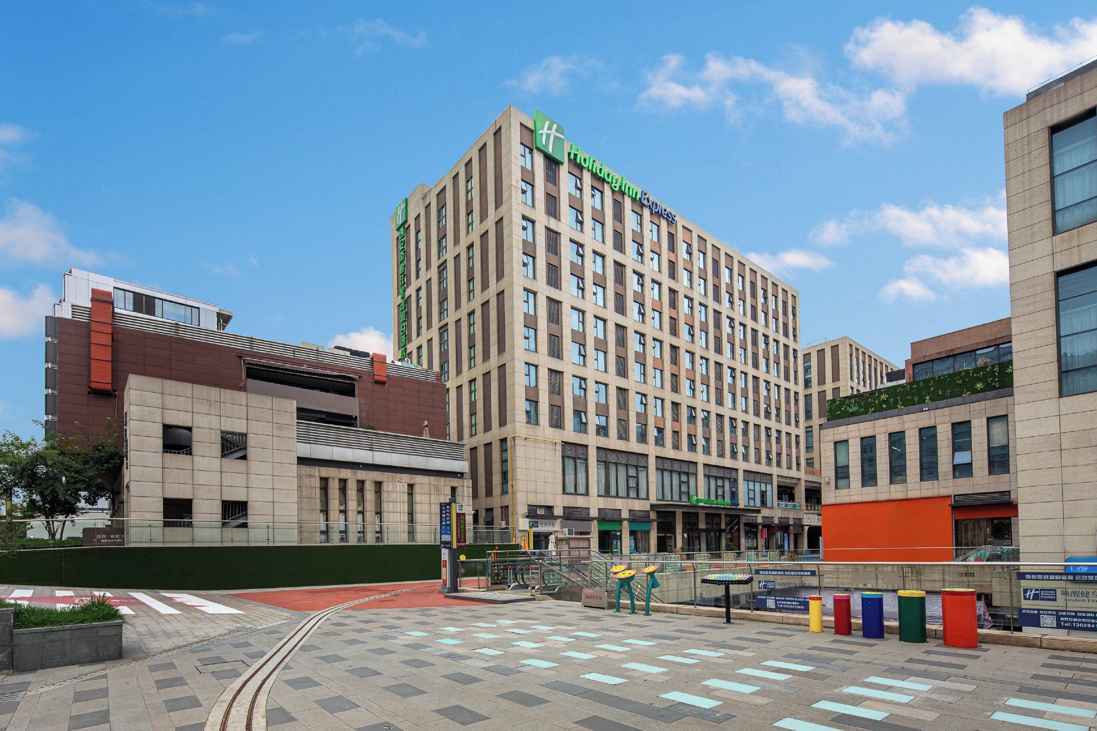 Un hotel moderno con un llamativo letrero verde, un patio central con un tranvía y diversos locales comerciales en la planta baja.