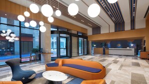 A modern lobby with a reception desk, seating areas, and large windows offering views of the street.