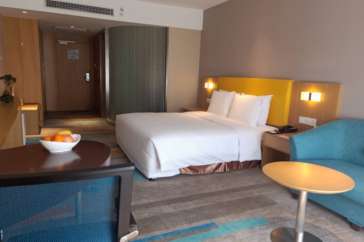 A hotel room with a bed, bedside table, chair, and a small table with a bowl of fruit.