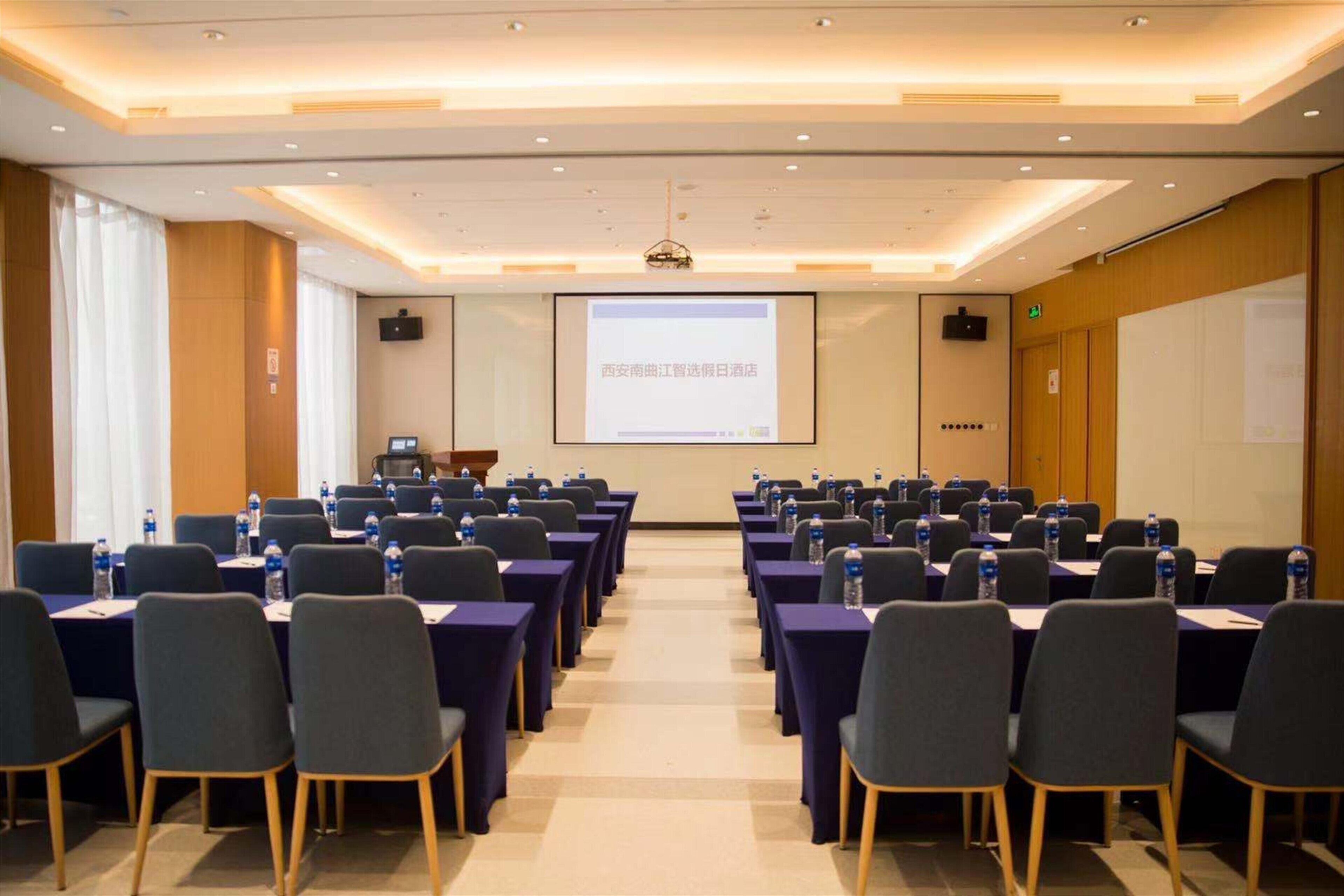 Un salón de conferencias con filas de mesas y sillas, una pantalla de proyector y una pantalla para presentaciones.