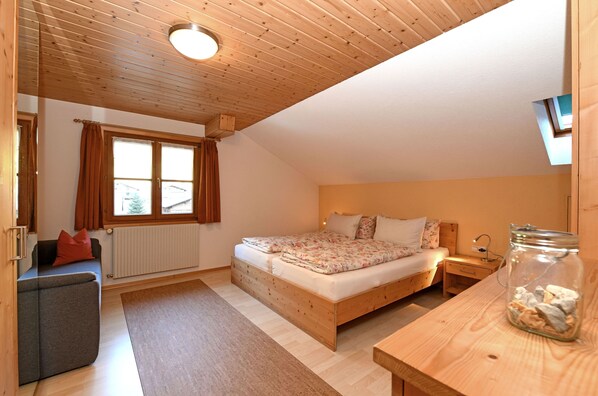 A bedroom with a wooden ceiling, a bed with floral bedding, a nightstand, a small table, and a window with curtains.