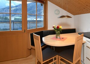 A dining area with a round table, wooden chairs, a bench, a vase with flowers, a clock on the wall, and a view of a lake and mountains through large windows.