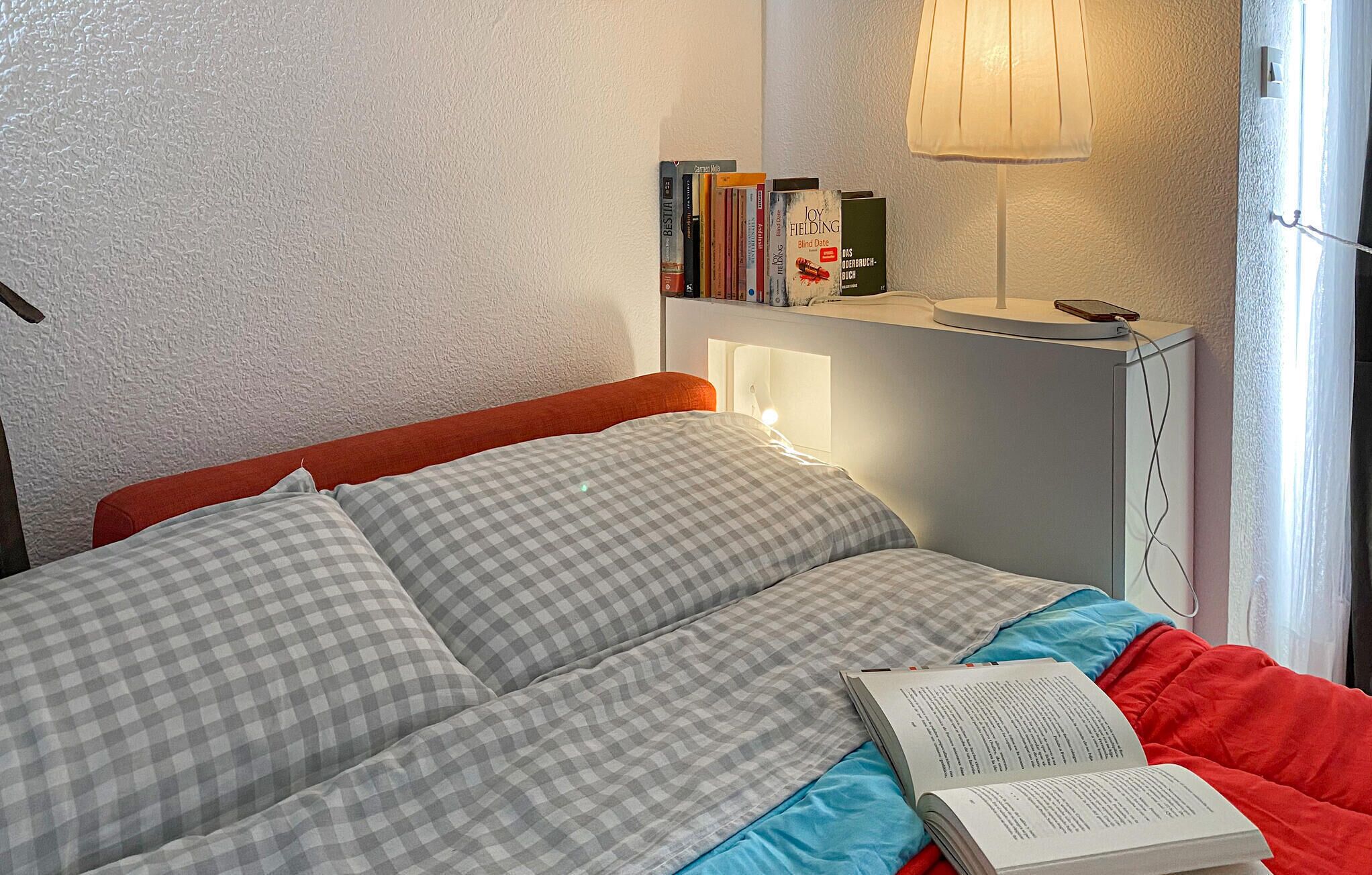 Ein Bett mit karierter Bettwäsche, eine Nachttischlampe, ein Nachttisch mit Büchern und ein Fenster mit Vorhängen.
