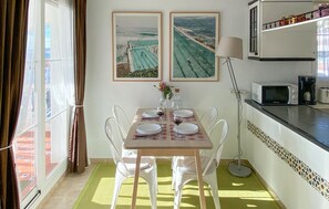 A dining area with a table set for four, two framed pictures on the wall, a lamp, and a kitchen area with a microwave and coffee maker.