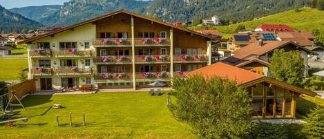 A multi-story hotel with balconies adorned with flowers, set against a mountainous backdrop.