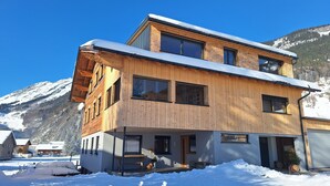 A modern two-story house with a wooden facade, large windows, and a snowy mountain backdrop. - Holiday at Rüf's - Apartment 1 (2-6 people) (Au)