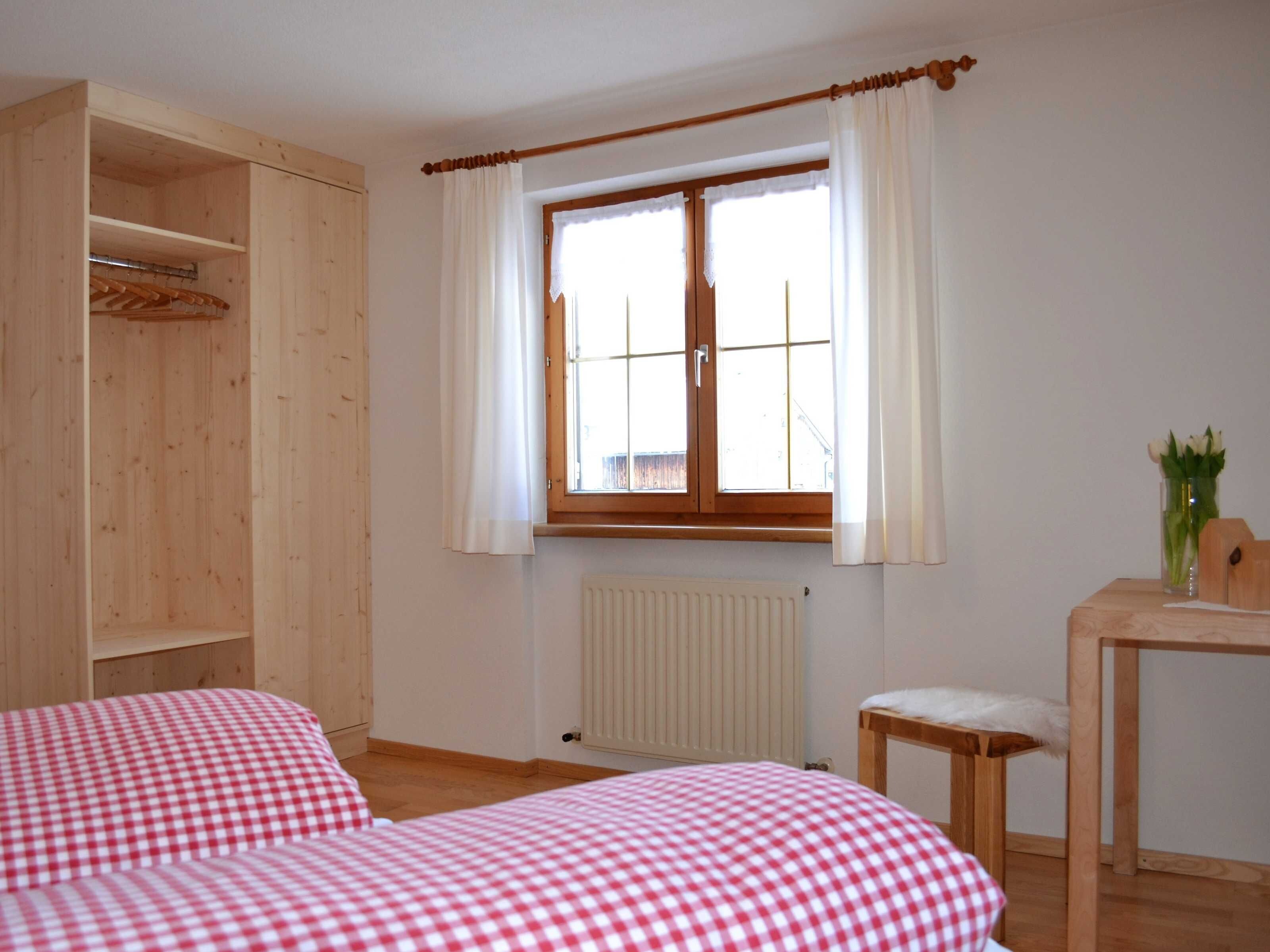A room with two beds, a wooden wardrobe, a window with curtains, a radiator, and a small table with a vase of flowers.