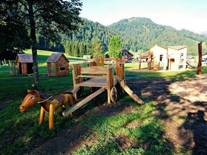 Un chariot en bois rustique, orné d’une figurine de cheval, entouré de petites constructions en bois et se dressant sur un fond de paysage montagneux.