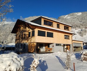 Une maison en bois à deux étages, dotée d’un porche abrité, entourée d’arbres et de montagnes couverts de neige.