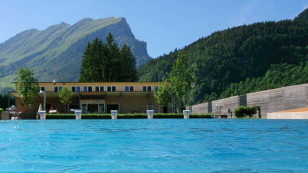 A swimming pool with a mountain backdrop, a building with multiple windows, and a wooden fence. - Holiday at Rüf's - Apartment 1 (2-6 people) (Au)