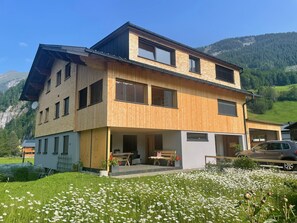 Une maison à deux étages avec un balcon, une voiture garée devant et un paysage montagneux en arrière-plan.