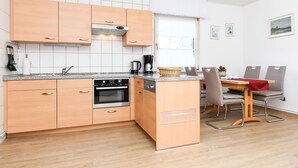 A compact kitchen with wooden cabinets, a built-in oven, and a dining area with a table and chairs. - Apartment Seerose - House Ursula (Esens)