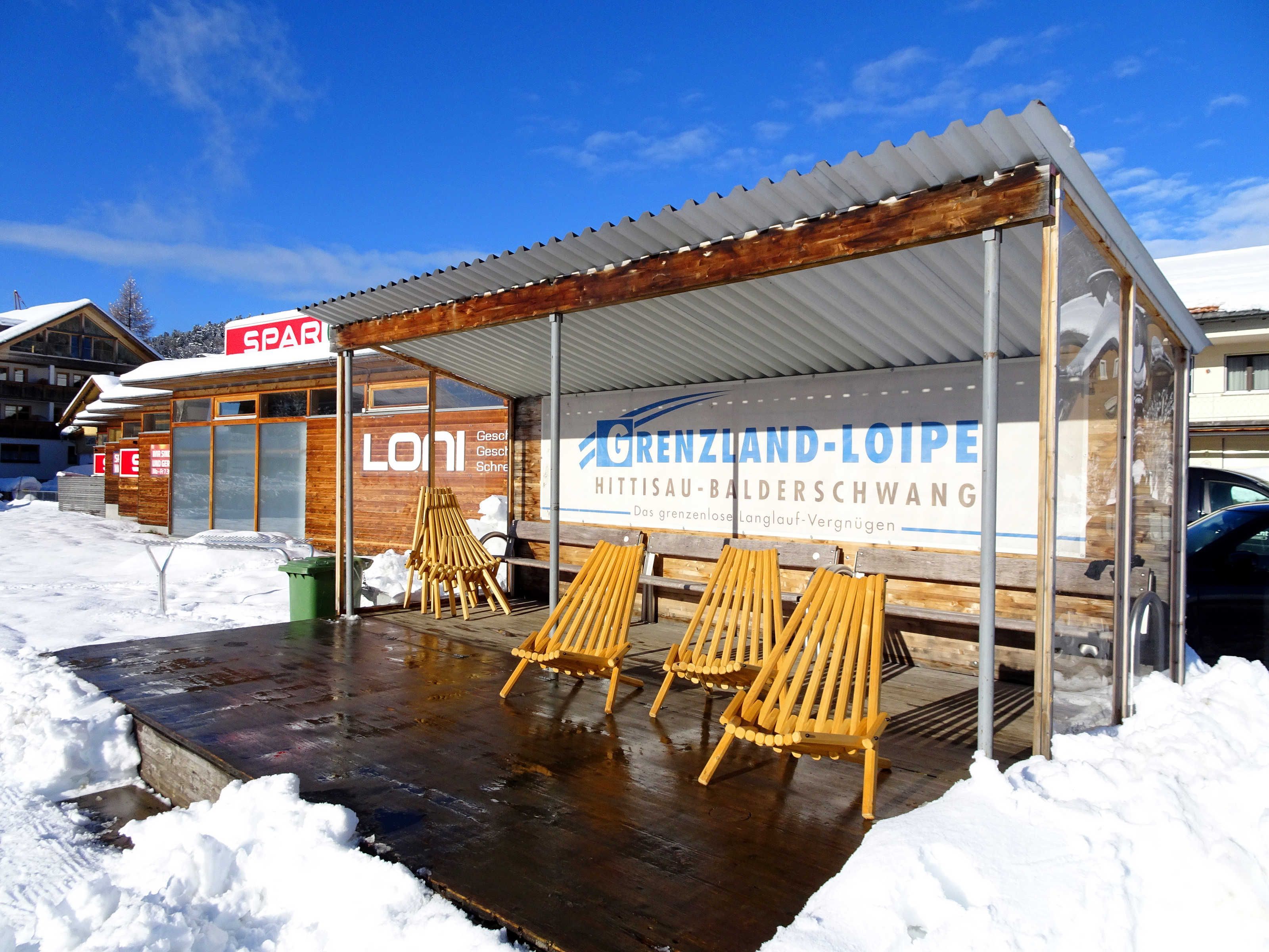 Een overdekt terras met houten stoelen, een houten gebouw met een bord en sneeuw op de grond.