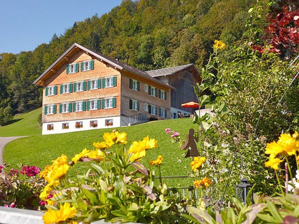Um edifício de vários andares com telhado inclinado, rodeado por um gramado bem cuidado e diversas flores.