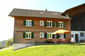 A two-story brick building with green shutters, a patio with chairs and an umbrella, and a wooden barn adjacent to it.