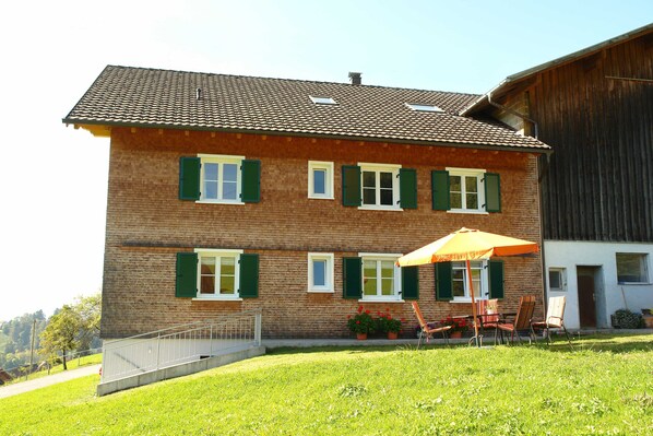 A two-story brick building with green shutters, a patio with chairs and an umbrella, and a wooden barn adjacent to it.