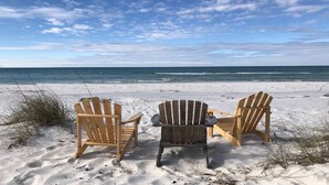 Two wooden Adirondack chairs facing the ocean on a sandy beach.