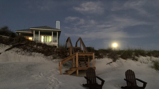 A beach house with a wooden staircase leading to a balcony, two Adirondack chairs, and a sandy beach.