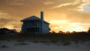 A two-story beach house with a prominent chimney, situated on a sandy beach with dunes in the background.