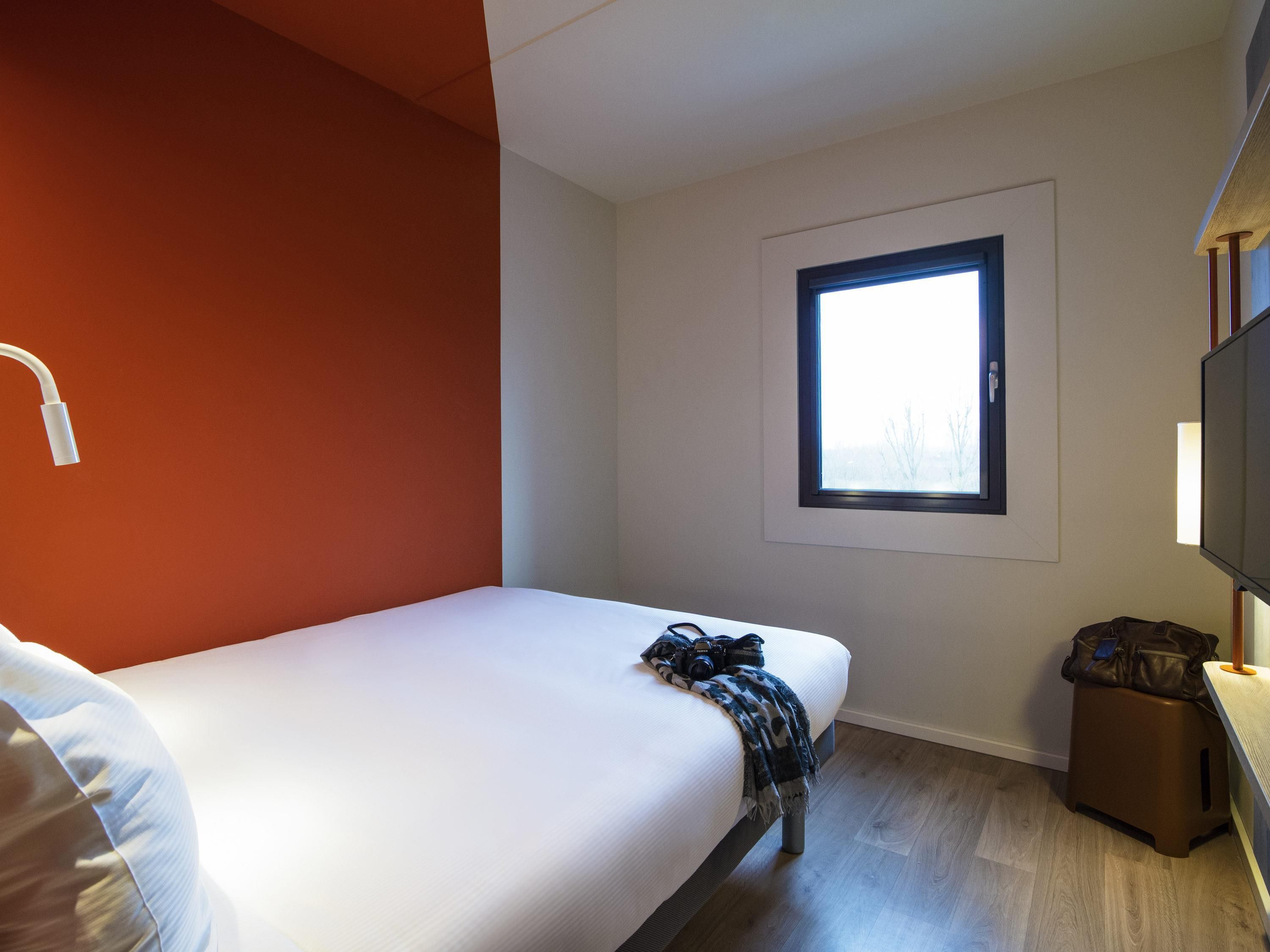 Een eenpersoonsbed met een witte matras, een klein nachtkastje met een lamp, een raam met uitzicht op bomen en een televisie aan de muur.