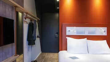 A hotel room with a bed, a TV, a coat rack, and a bag on the floor.