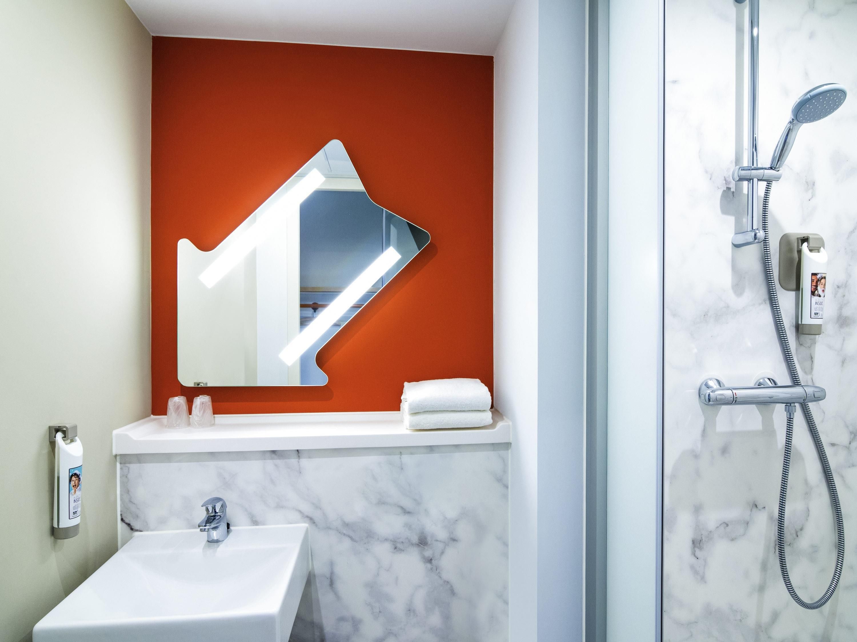 Ein Badezimmer mit einem weißen Waschbecken, einem Spiegel an einer roten Akzentwand und einer Dusche mit Duschbrause.