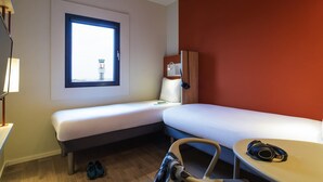 A small hotel room with a single bed, a desk, a chair, a window with a view of a tower, and a red accent wall. - Ibis budget Stein Maastricht (Stein)