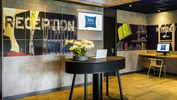 A reception area with a laptop on a table, a potted plant, and a screen displaying 'ibis budget hotels'. - Ibis budget Stein Maastricht (Stein)