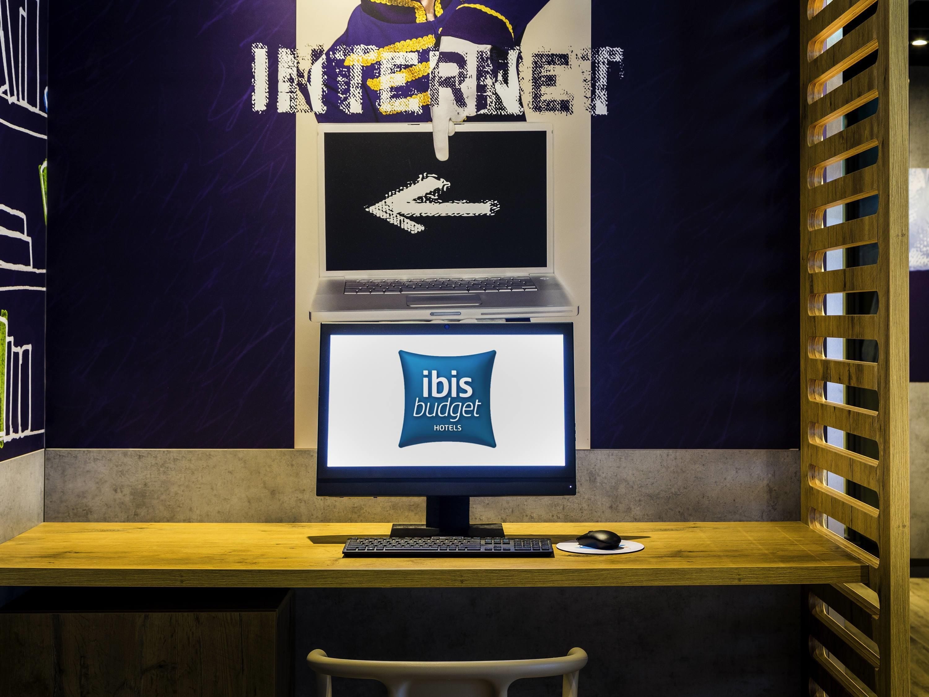 A workspace with a laptop, keyboard, and mouse on a wooden desk, a computer monitor displaying 'ibis budget hotels,' and a wall with a large 'INTERNET' sign and an arrow pointing left.
