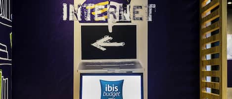A workspace with a laptop, keyboard, and mouse on a wooden desk, a computer monitor displaying 'ibis budget hotels,' and a wall with a large 'INTERNET' sign and an arrow pointing left.